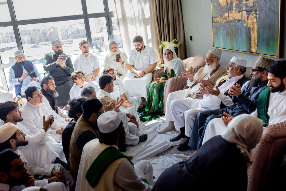 A group of people dressed in white and traditional clothing gather for a cultural or religious gathering in a sunlit room.