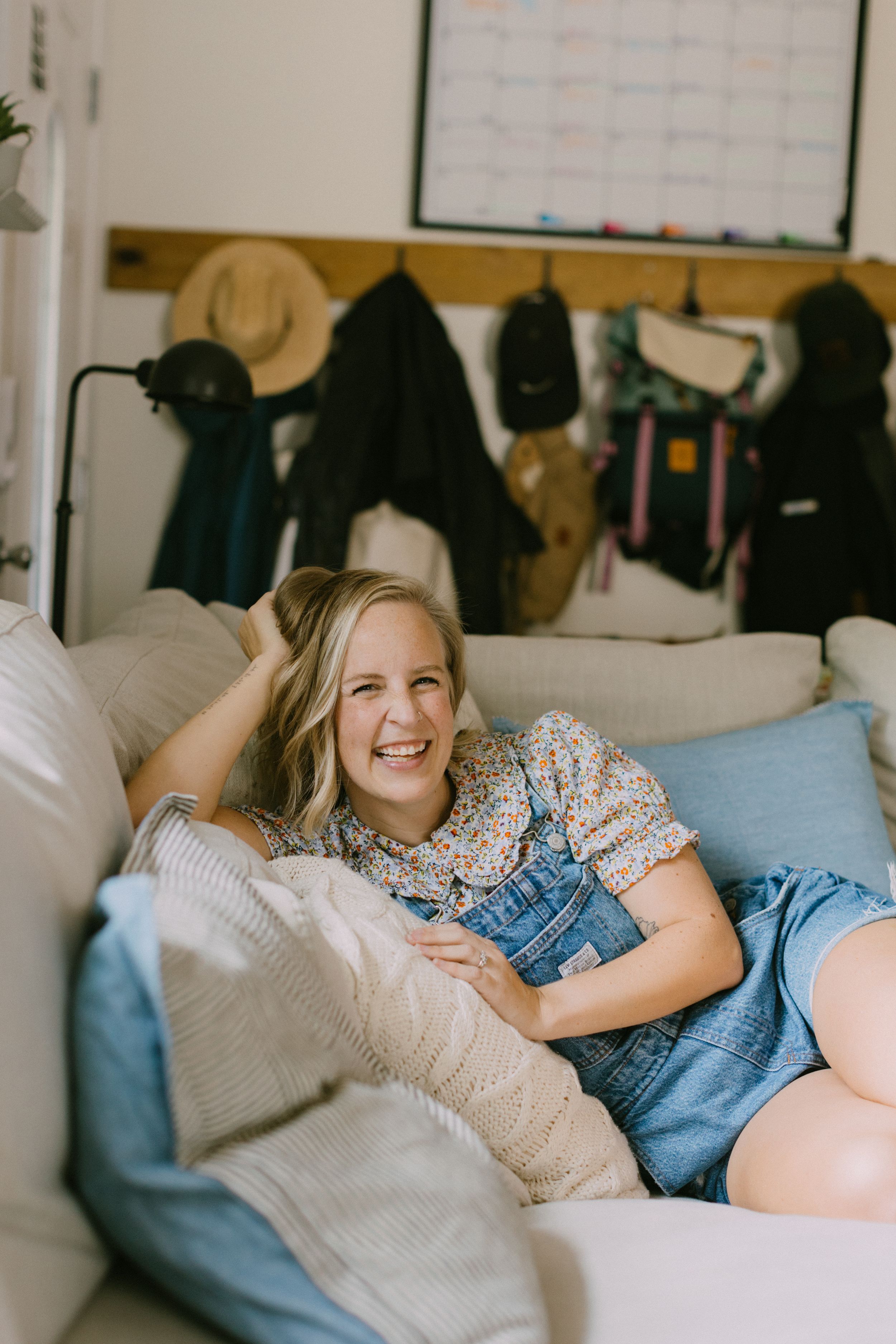 A girl relaxes on a comfortable couch wearing denim overalls and a floral top. She is happy and smiling at the camera.