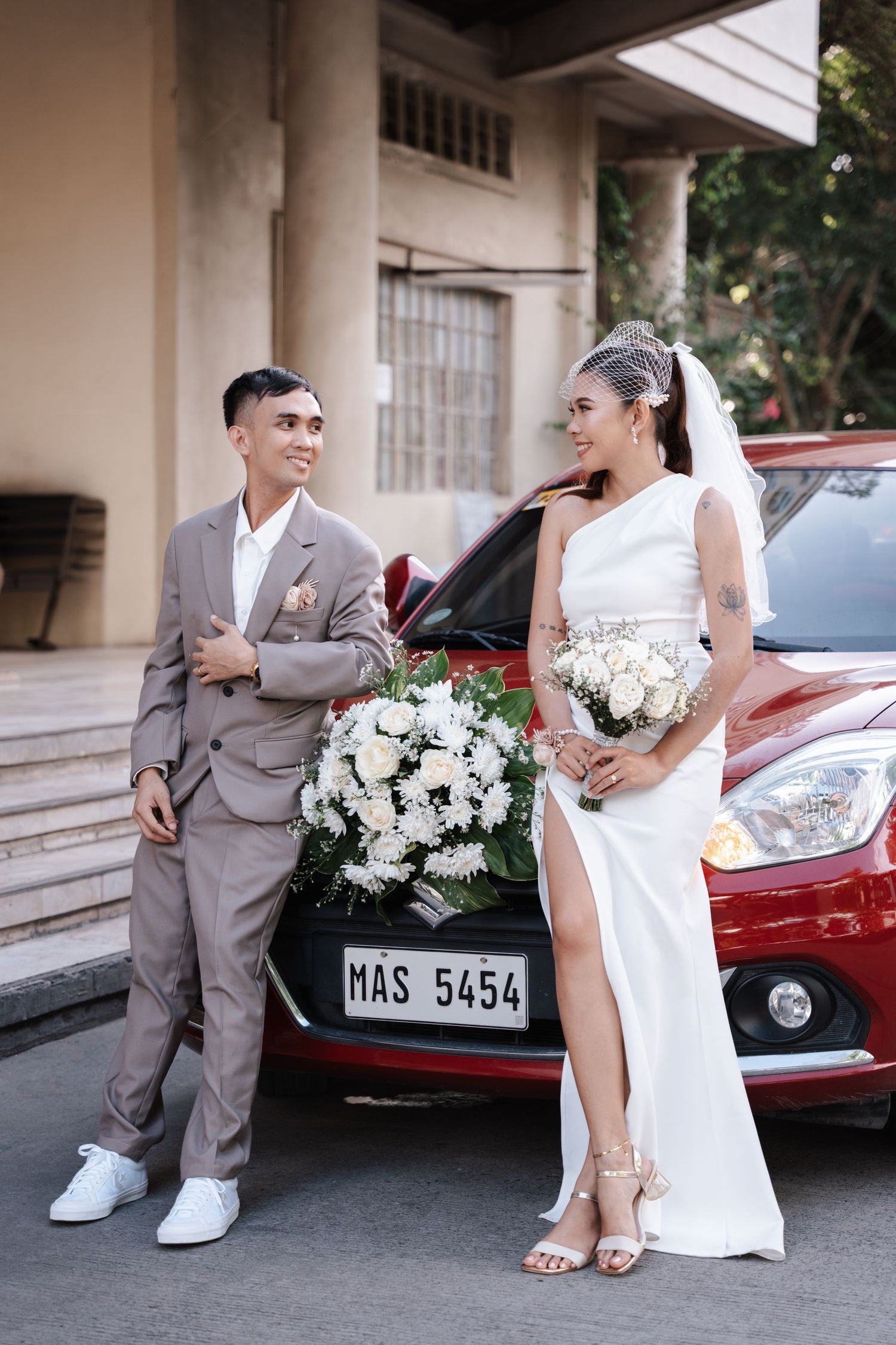 Wedding couple posing next to red car, bride in white dress and groom in grey suit.