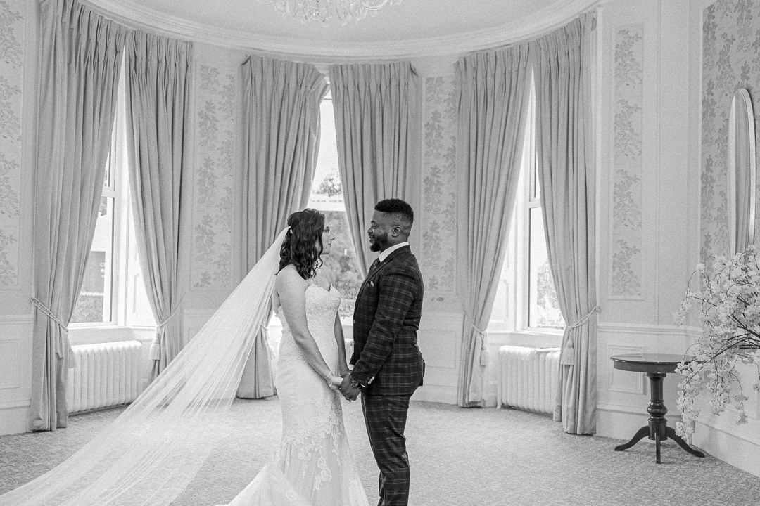 A black and white wedding photo of a couple standing in an elegant room with tall windows and drapes.