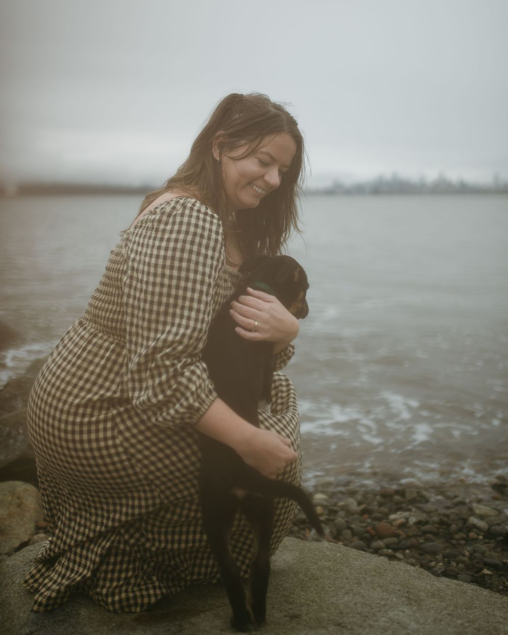 Woman in checkered dress hugging a black dog while sitting on rocks by the water on foggy day.