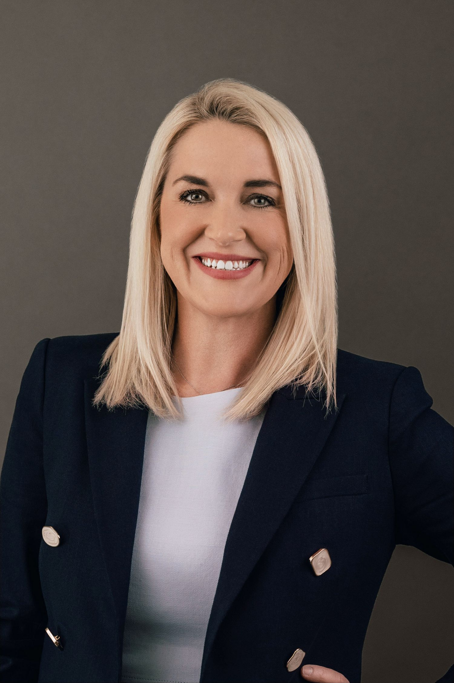 Professional headshot of a person with blonde hair wearing a navy blazer and white top, smiling against a dark background.