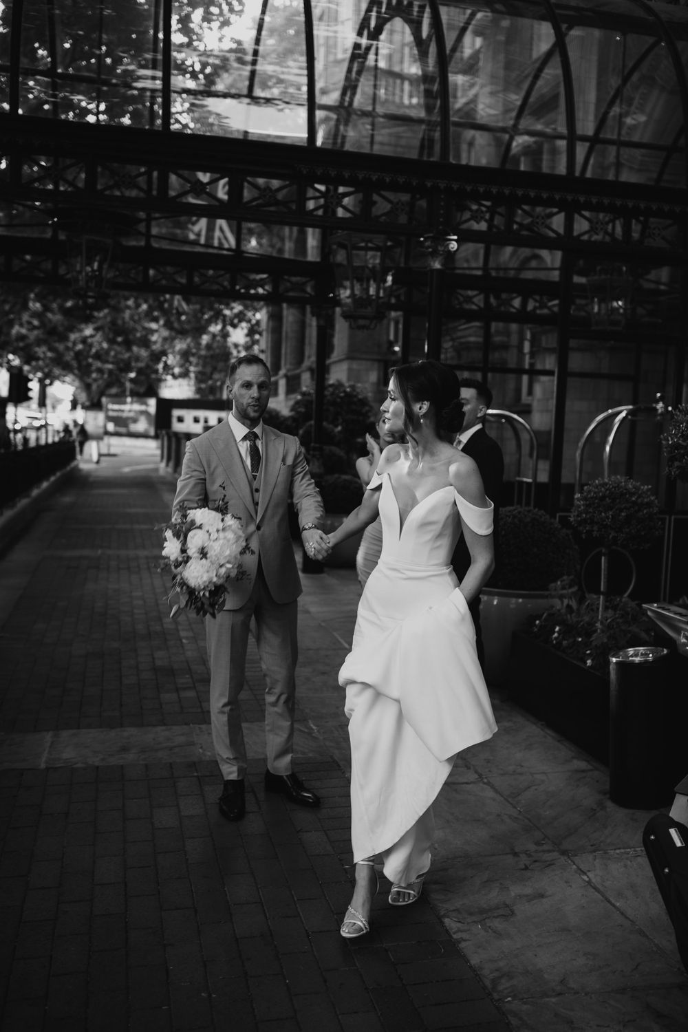 A couple in wedding attire walks together on a sidewalk under an architectural overhang in black and white photography.