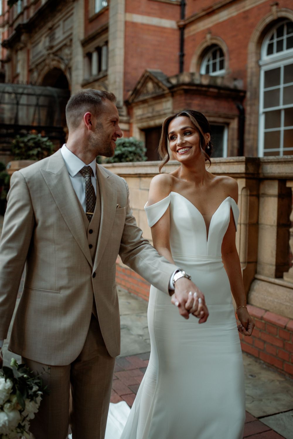 A bride in an elegant off-shoulder white gown and groom in beige suit pose together outside a historic brick building.