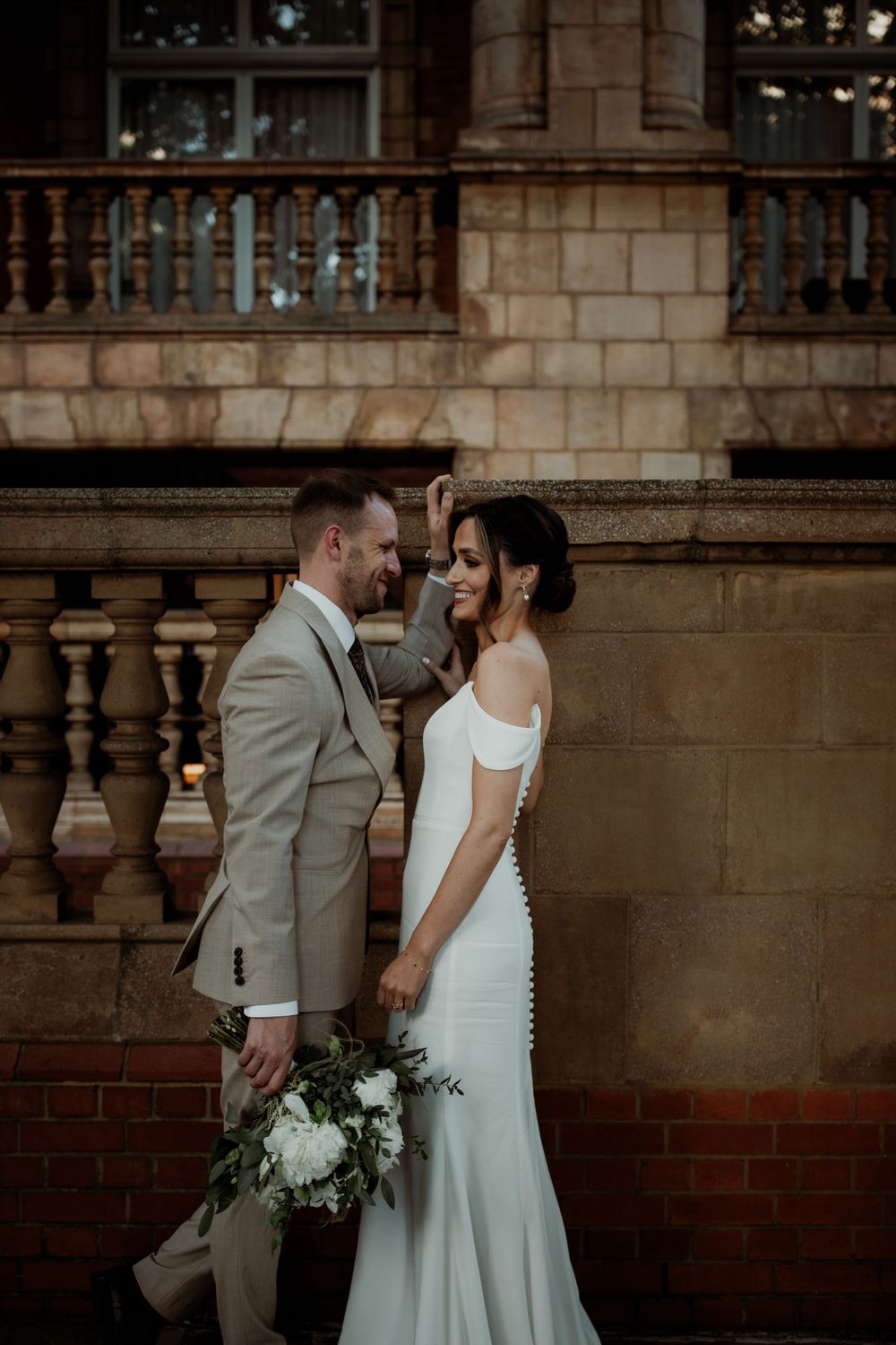 A romantic wedding moment captured against a stone balustrade wall with white florals and elegant bridal attire.