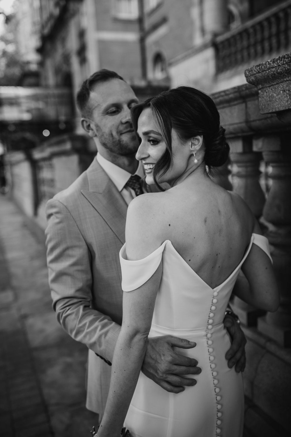Black and white wedding photo showing intimate moment between newlyweds on city street, bride wearing elegant backless dress.