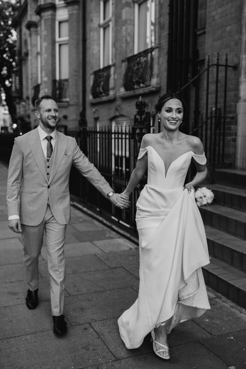 Black and white wedding photo showing a couple walking hand in hand down a city street, with bride in flowing white gown.