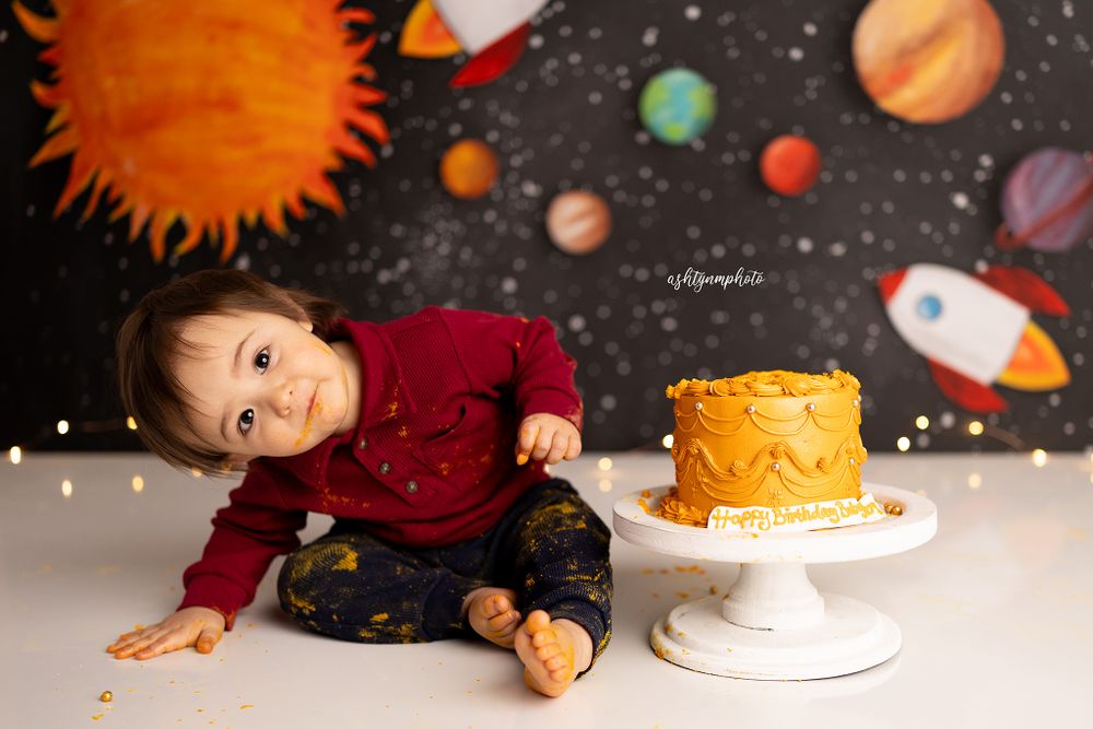 Space-themed cake smash photo setup with yellow cake on white stand and colorful planets painted on dark backdrop.