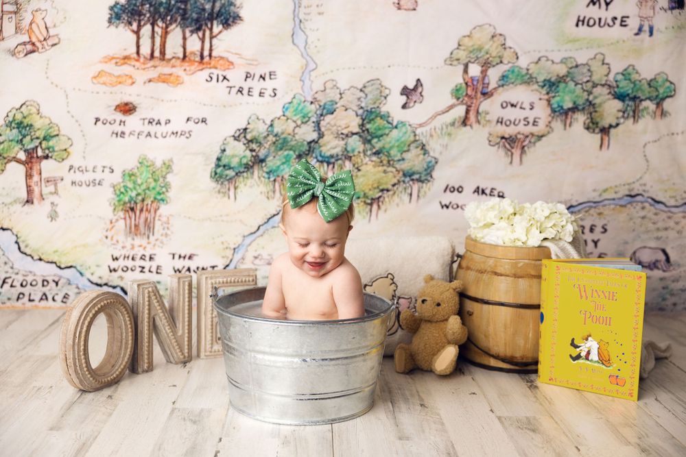 A baby wearing a green leaf headpiece sits in a galvanized tub with rustic decor and woodland-themed backdrop.
