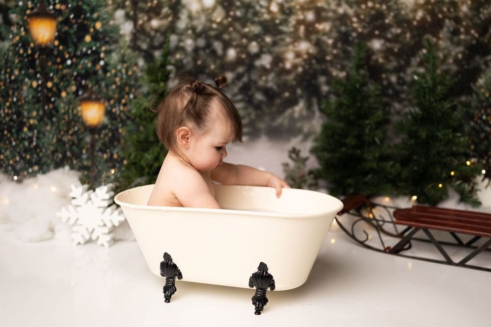 Baby bathing in vintage clawfoot tub surrounded by snow and pine trees with twinkling lights in holiday photography setup.