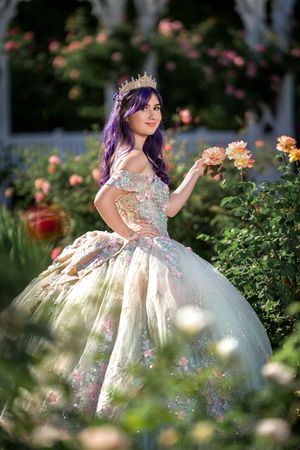 A person in a floral embellished ball gown poses elegantly in a garden setting with blooming roses.