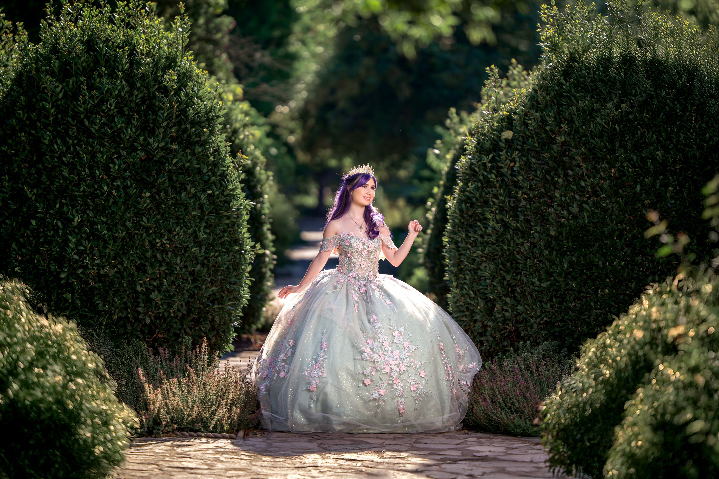 A person in a pink ball gown poses elegantly in a lush garden setting with manicured hedges and greenery.