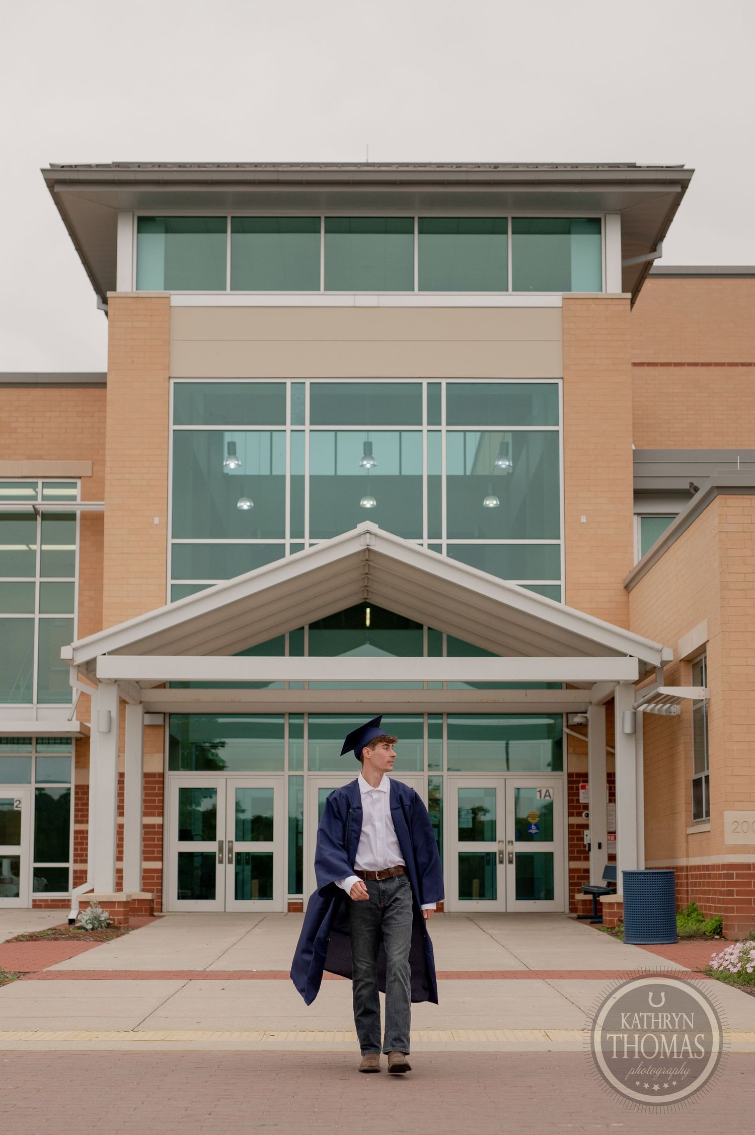Graduate walking confidently in cap and gown outside school building entrance.