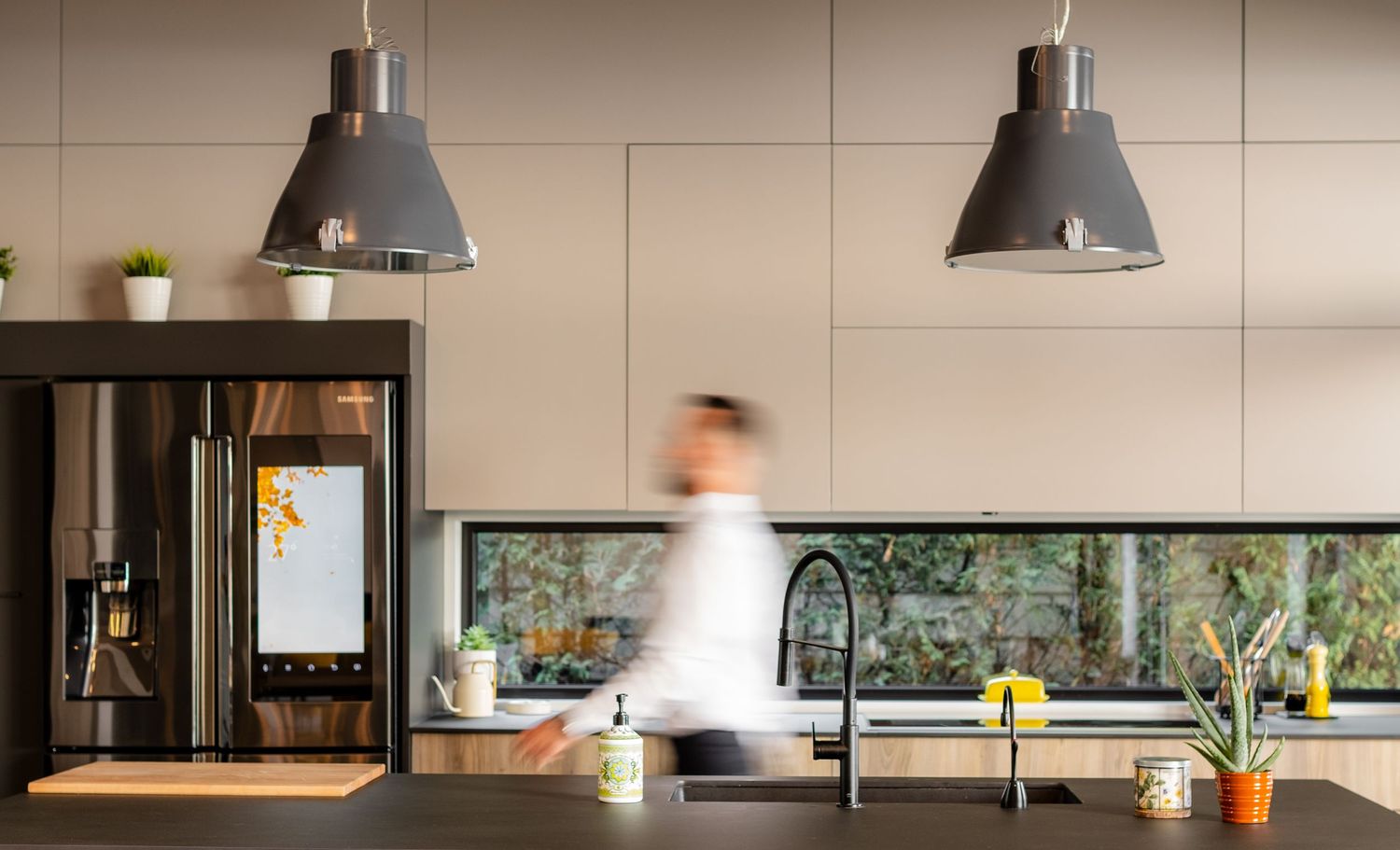 Modern kitchen interior with pendant lamps, blurred motion figure, and horizontal window view overlooking greenery.