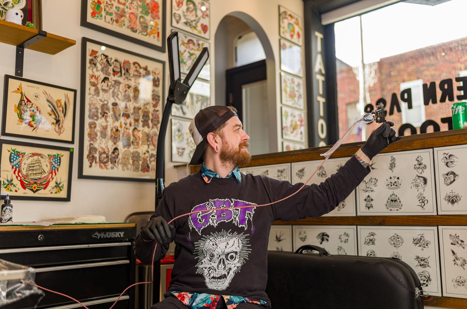 Tattoo artist points to traditional flash artwork displayed on walls inside vintage-style tattoo shop.