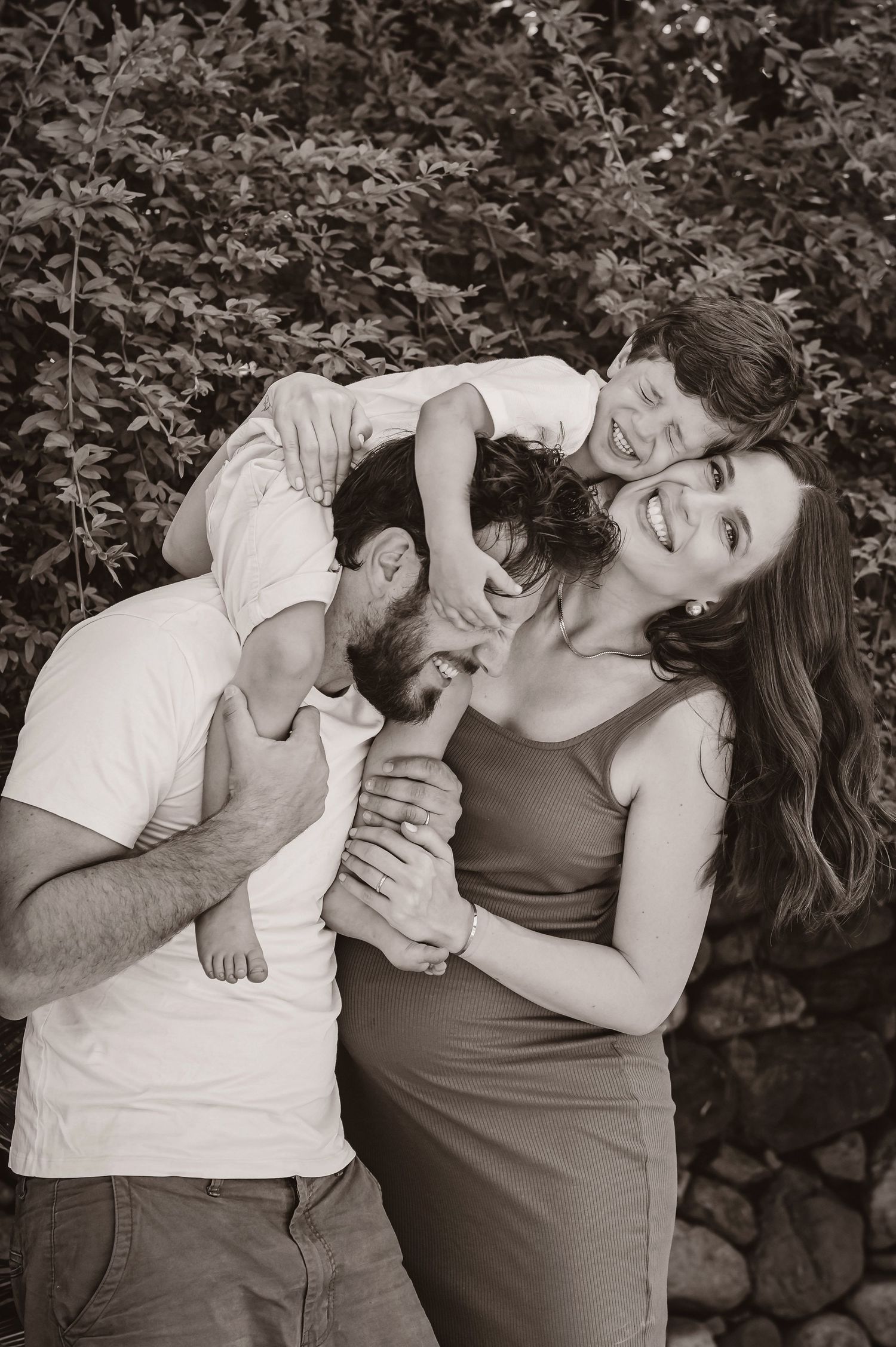 A black and white family photo showing joyful candid moment outdoors with natural foliage in background.