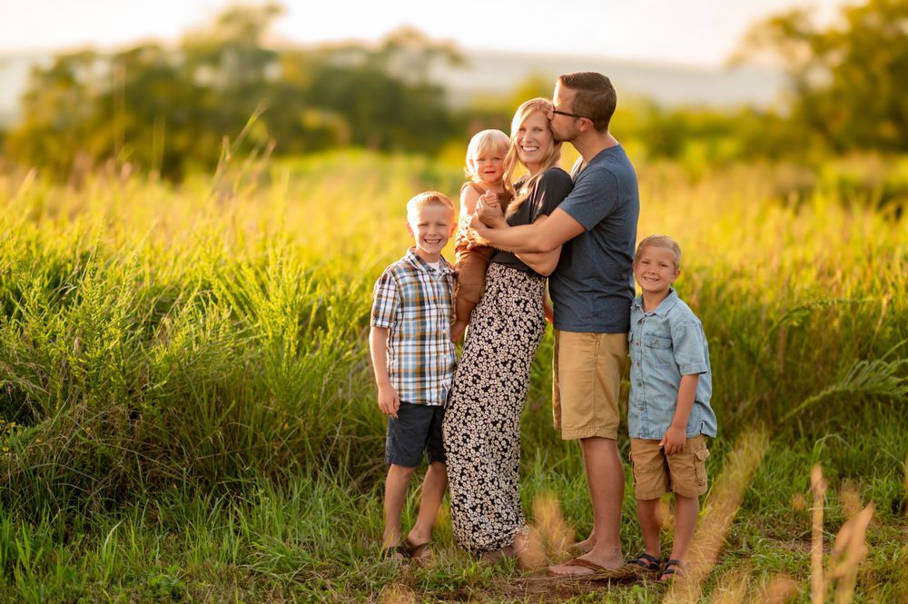A loving family embraces together on a hill at golden hour in a tall grass meadow during summer.