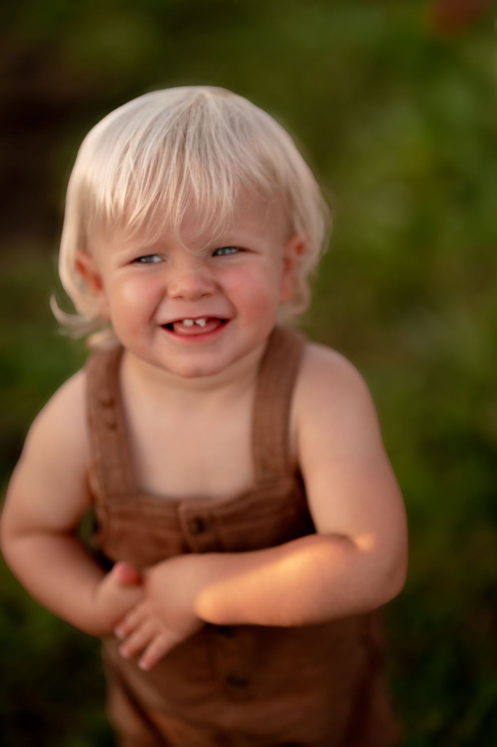 A cheerful young toddler in brown overalls playing outdoors on a sunny day with green background.