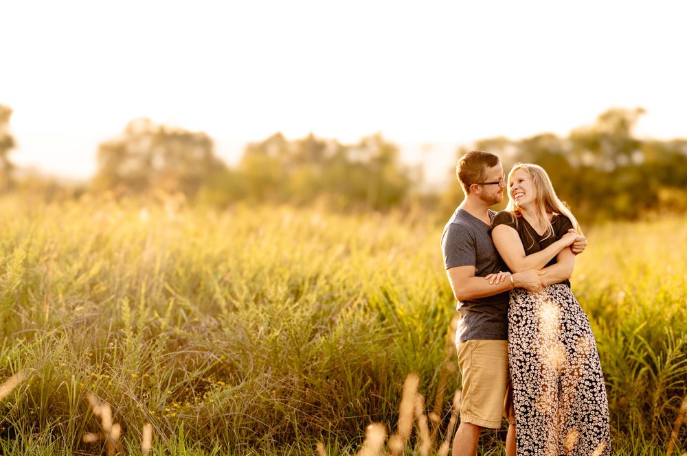 A couple embracing in a golden sunlit meadow at sunset with tall grass and trees in the background.