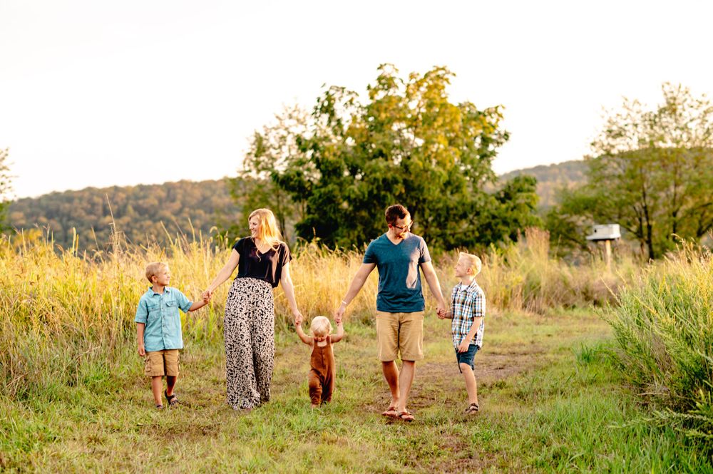 A family holds hands while walking together through a sunny meadow with trees in the background during golden hour.