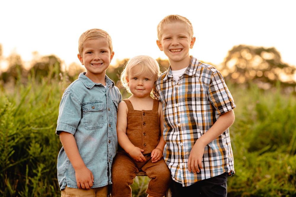 Three children in casual clothing pose together outdoors at sunset in a grassy field.
