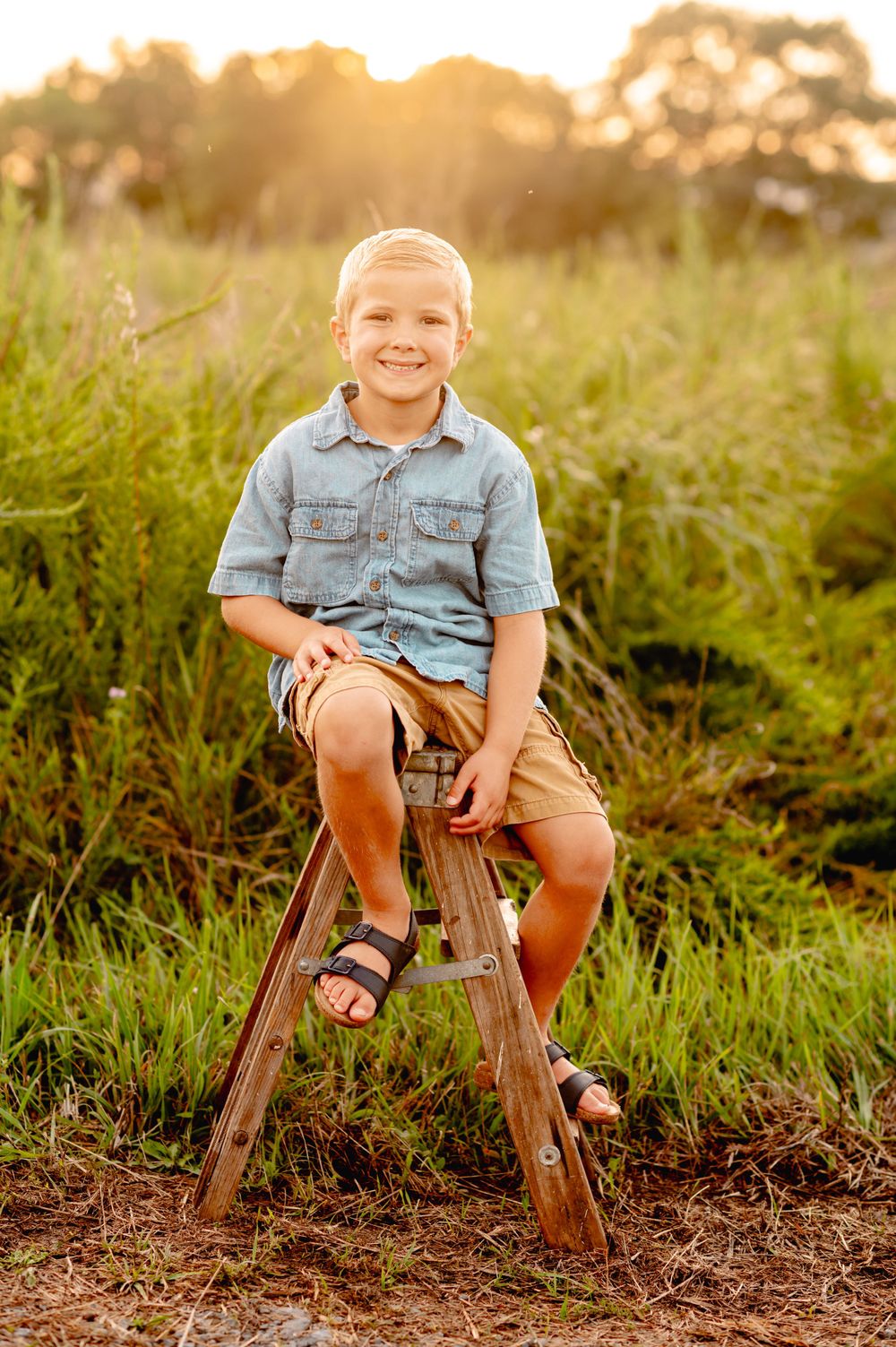 Child sitting on wooden stool in meadow wearing denim shirt and khaki shorts at golden hour.