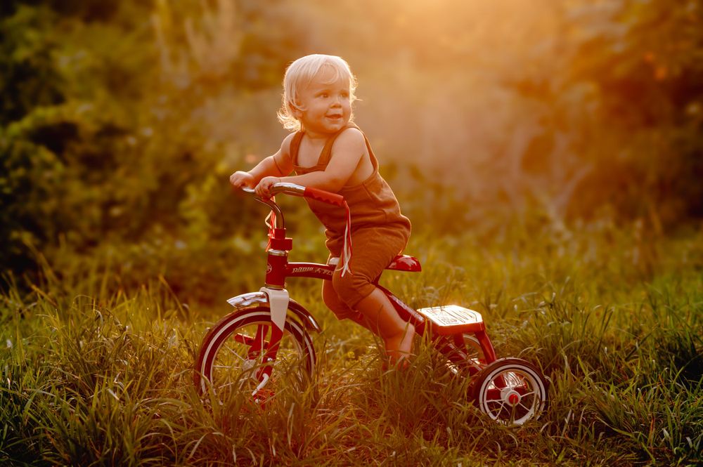 Small child rides a red tricycle on grassy terrain during golden hour sunset lighting.