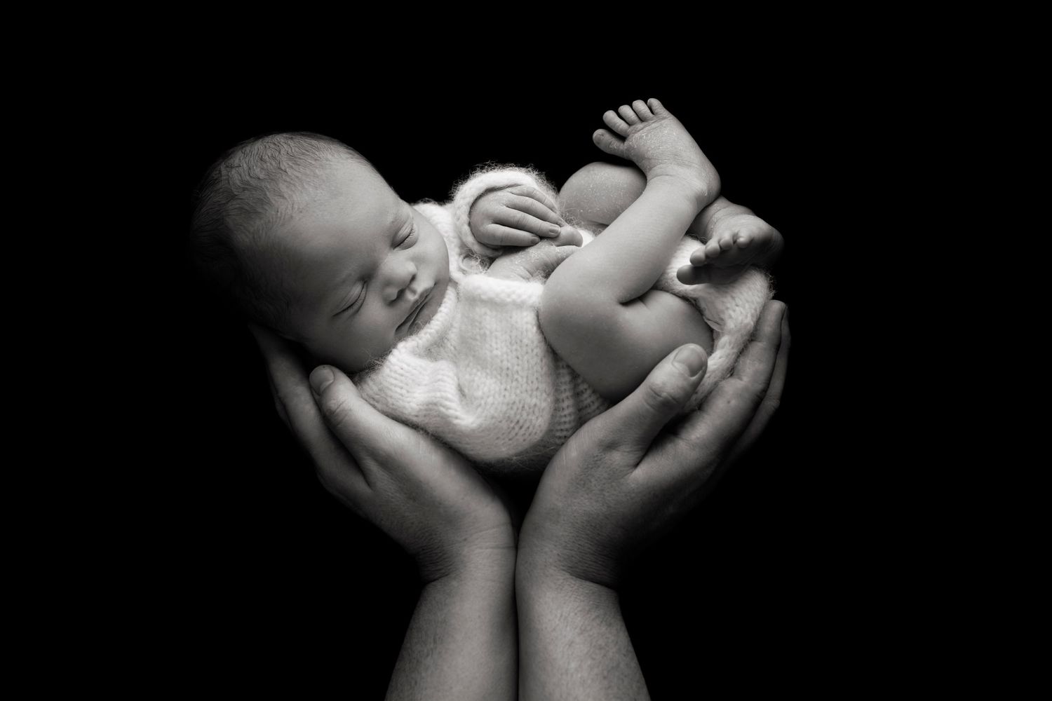 A black and white photograph of a tiny newborn baby cradled in his father's hands against a dark background.