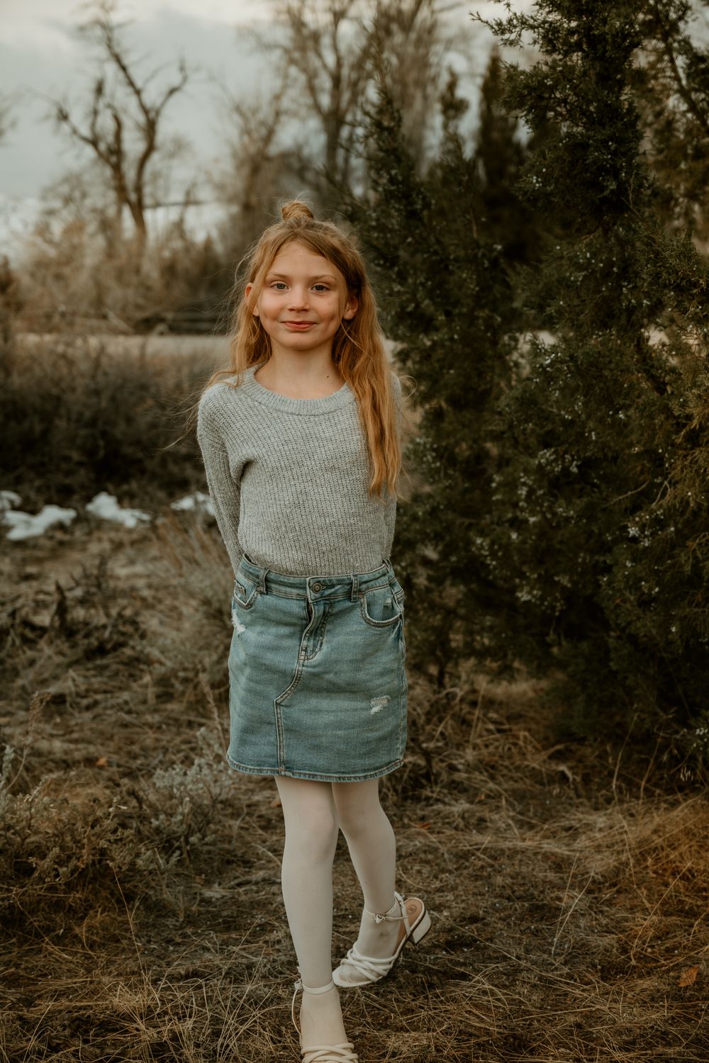 Child in gray sweater and denim skirt posing outdoors in natural setting.