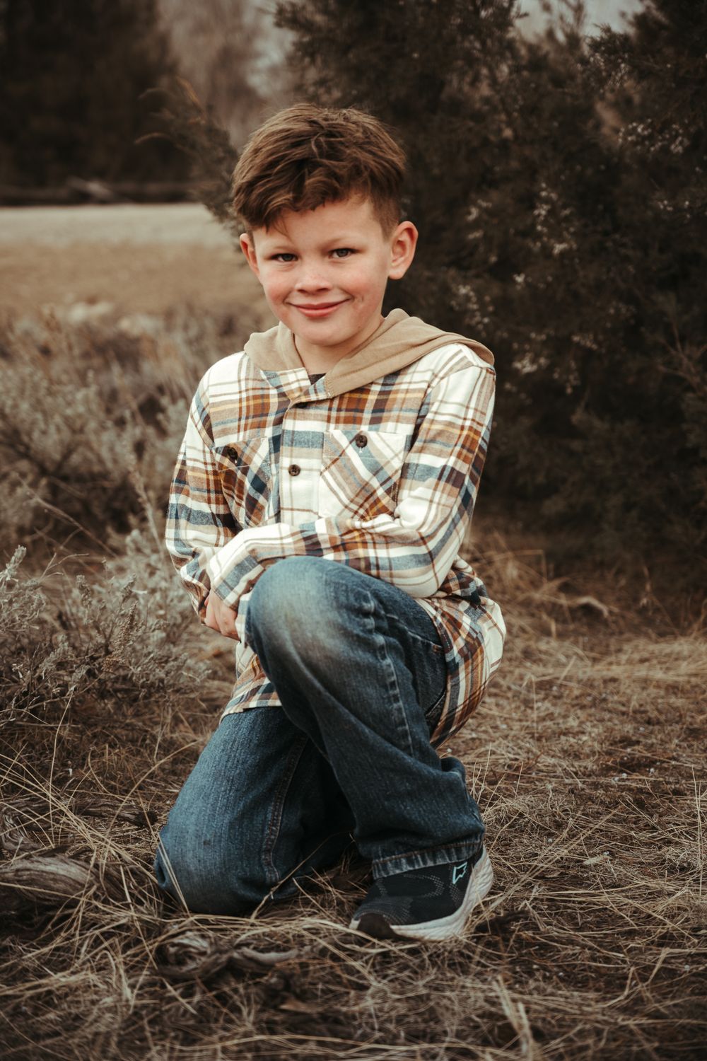 Young child in plaid shirt and jeans poses playfully on ground during outdoor photoshoot.