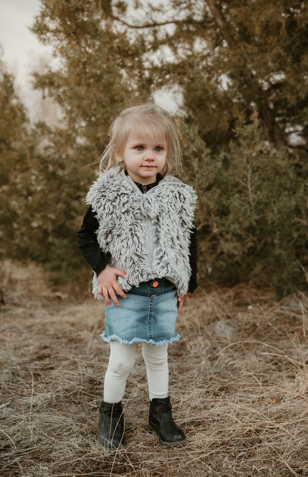 Small child wearing fuzzy vest and denim poses outdoors in natural setting.