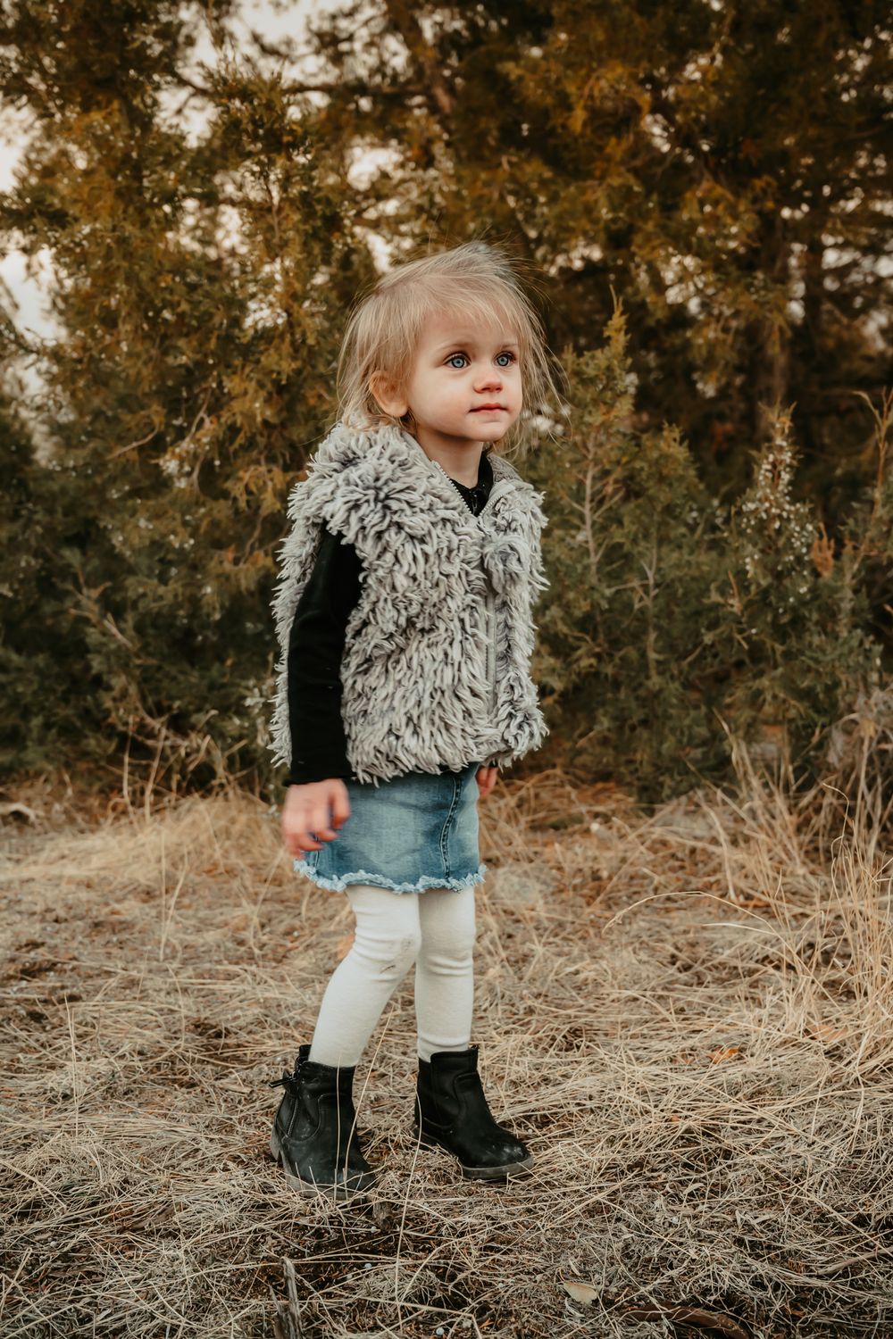 Young child in shaggy vest and leggings stands in rustic outdoor setting.