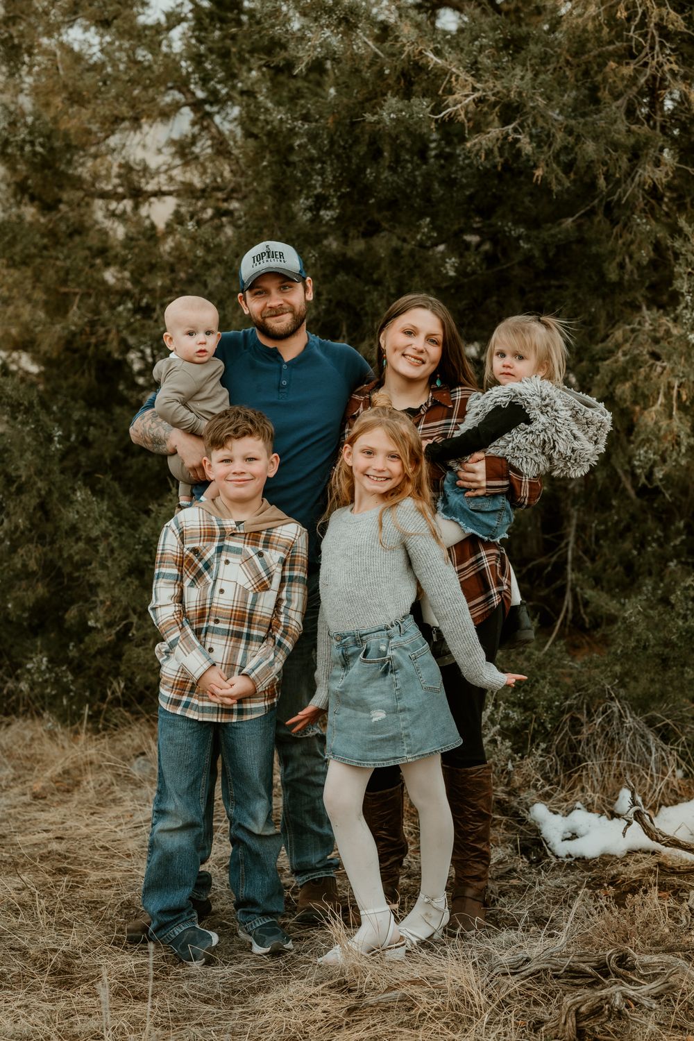 Family group posing together outdoors against evergreen trees in casual attire.