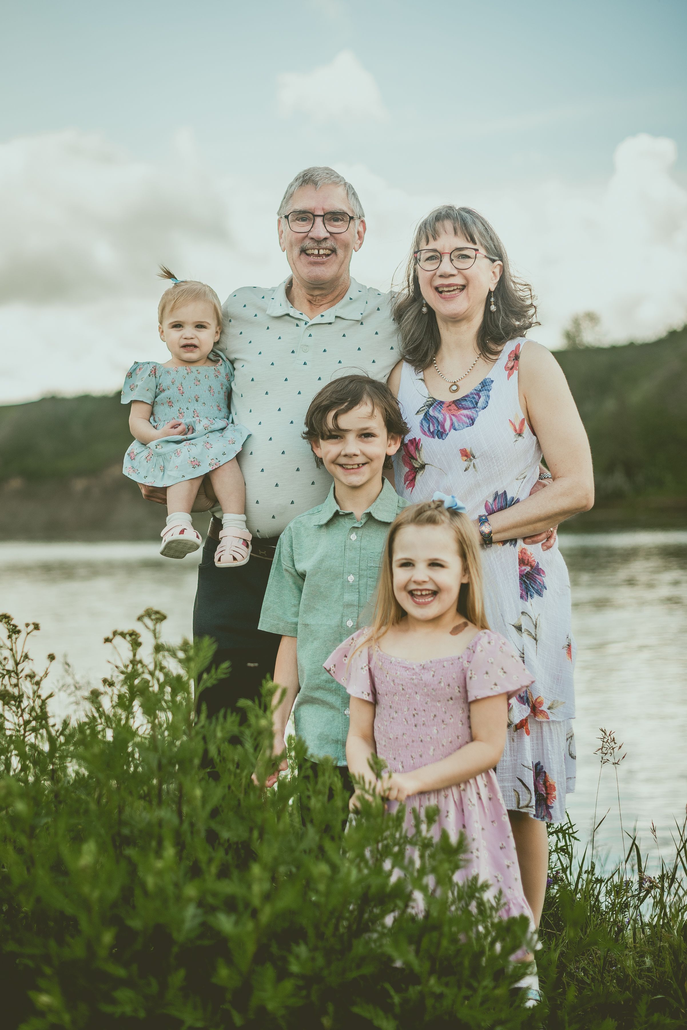 A loving family poses together by a lake on a cloudy day wearing coordinated summer outfits.