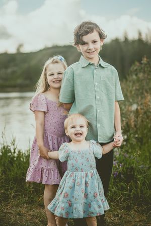 Three children stand together outdoors by a lake wearing pastel colored summer dresses and shirts.