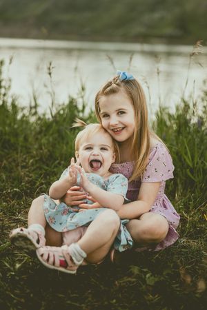 Two young children sitting in grass by a lake, hugging and laughing joyfully on a sunny day.