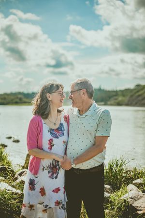 Happy elderly couple sharing laughter and embracing by a scenic waterfront on a bright, cloudy day.