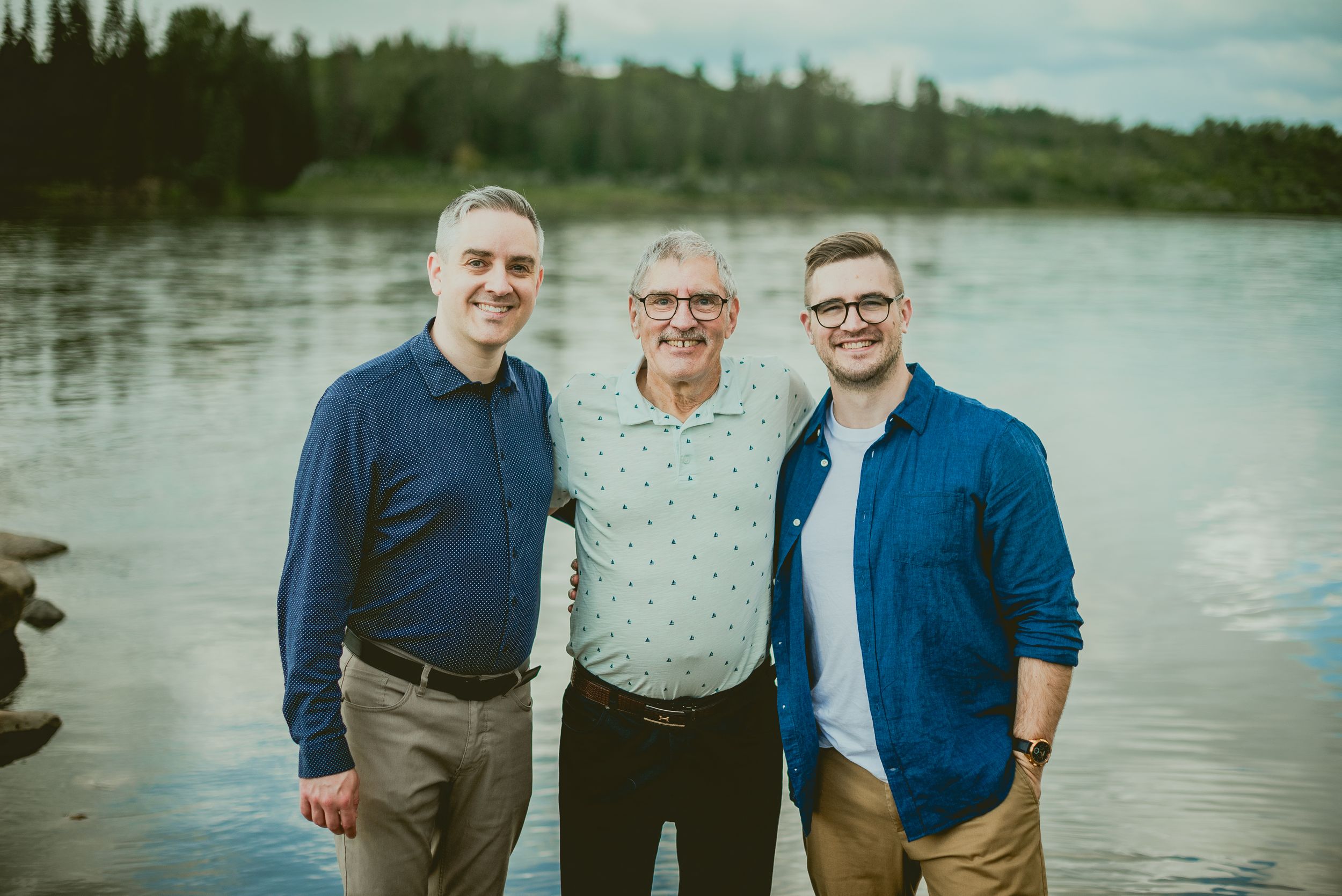 Three generations of men smiling together on a lakeshore with evergreen trees in the background.