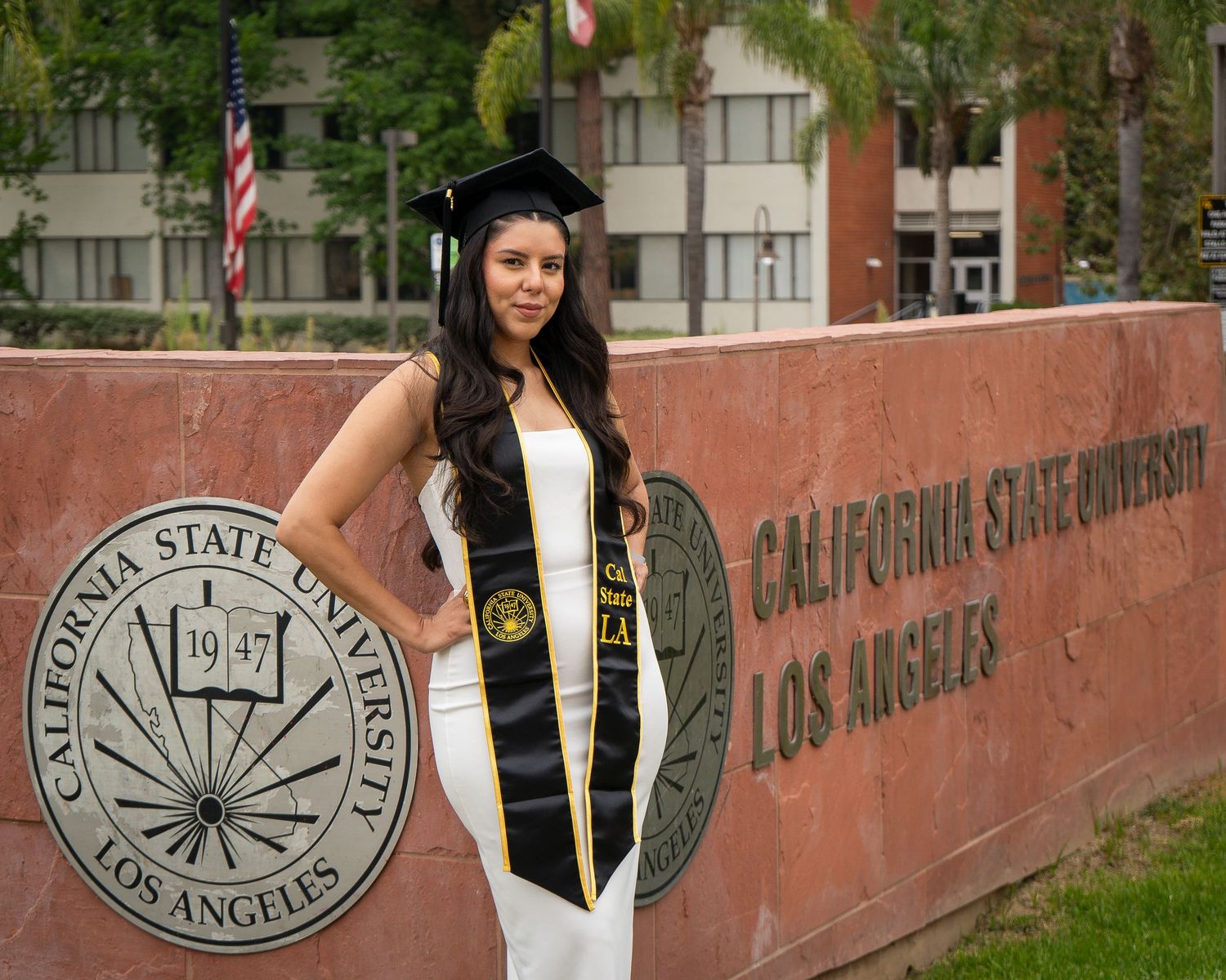 Graduate in white dress and stole standing by a “California State University Los Angeles” campus sign.