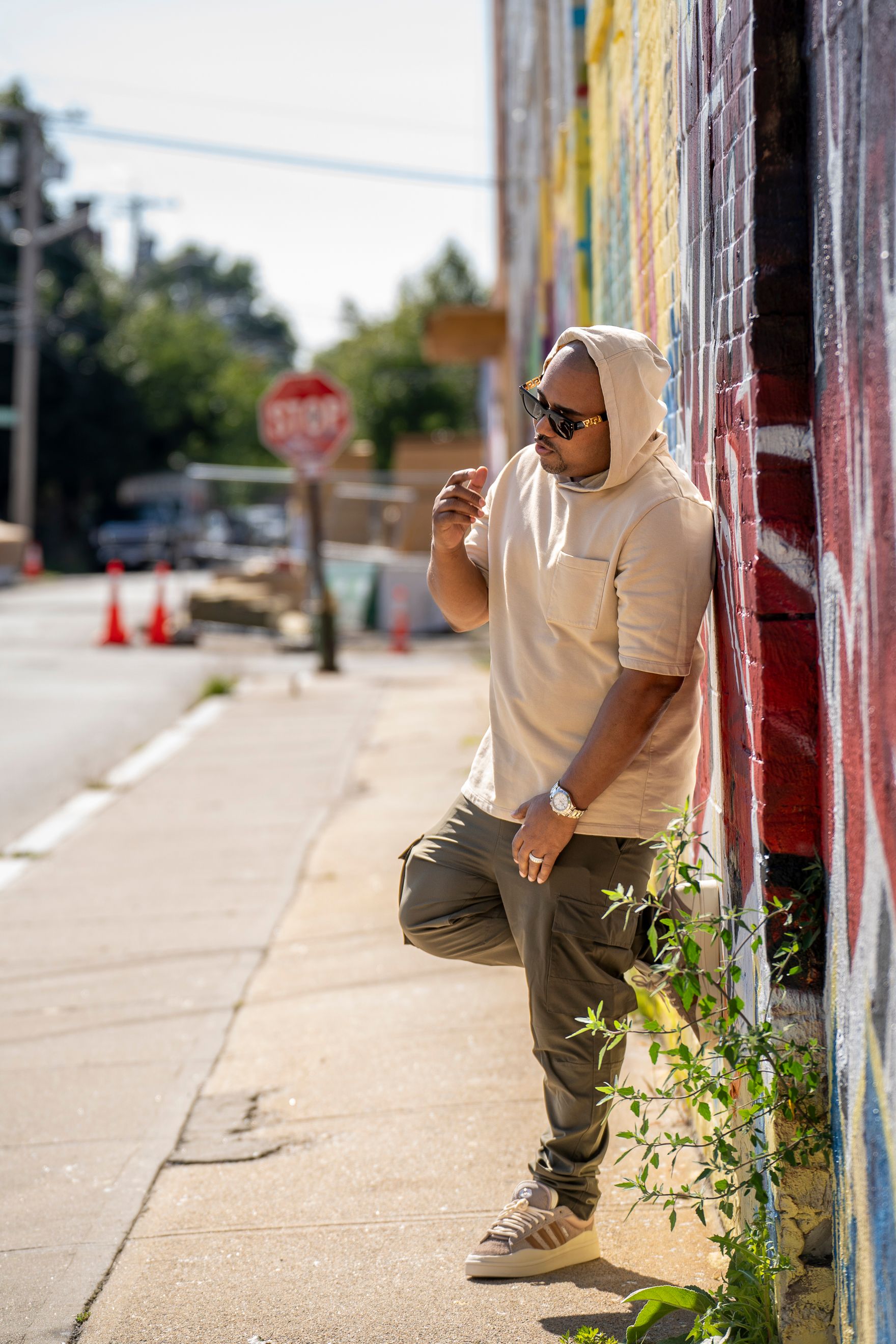 Person in a beige hoodie and sunglasses leaning against colorful wall on urban sidewalk with traffic cones in background.