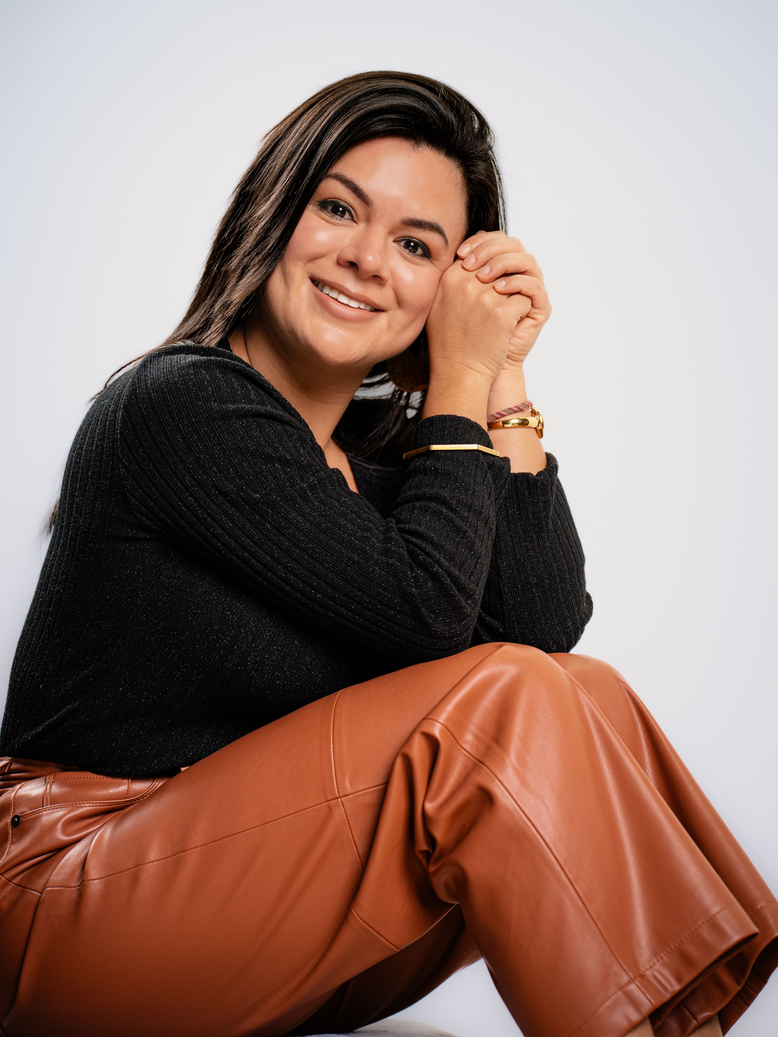 A woman in a black sweater and brown leather-look pants poses in a contemporary studio setting for fashion photos.