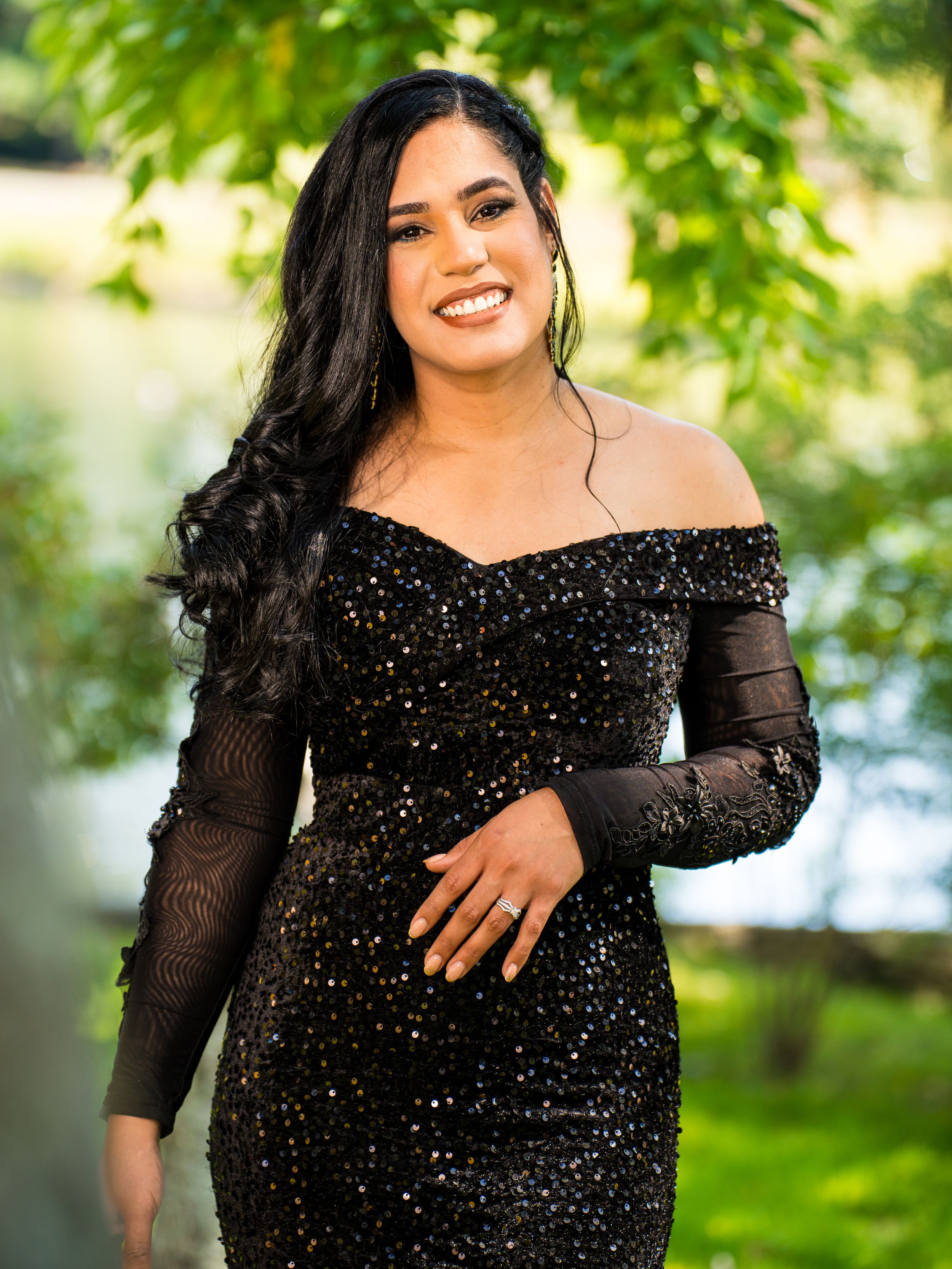 A person in a sparkly black off-shoulder evening gown poses outdoors against green foliage in natural lighting.