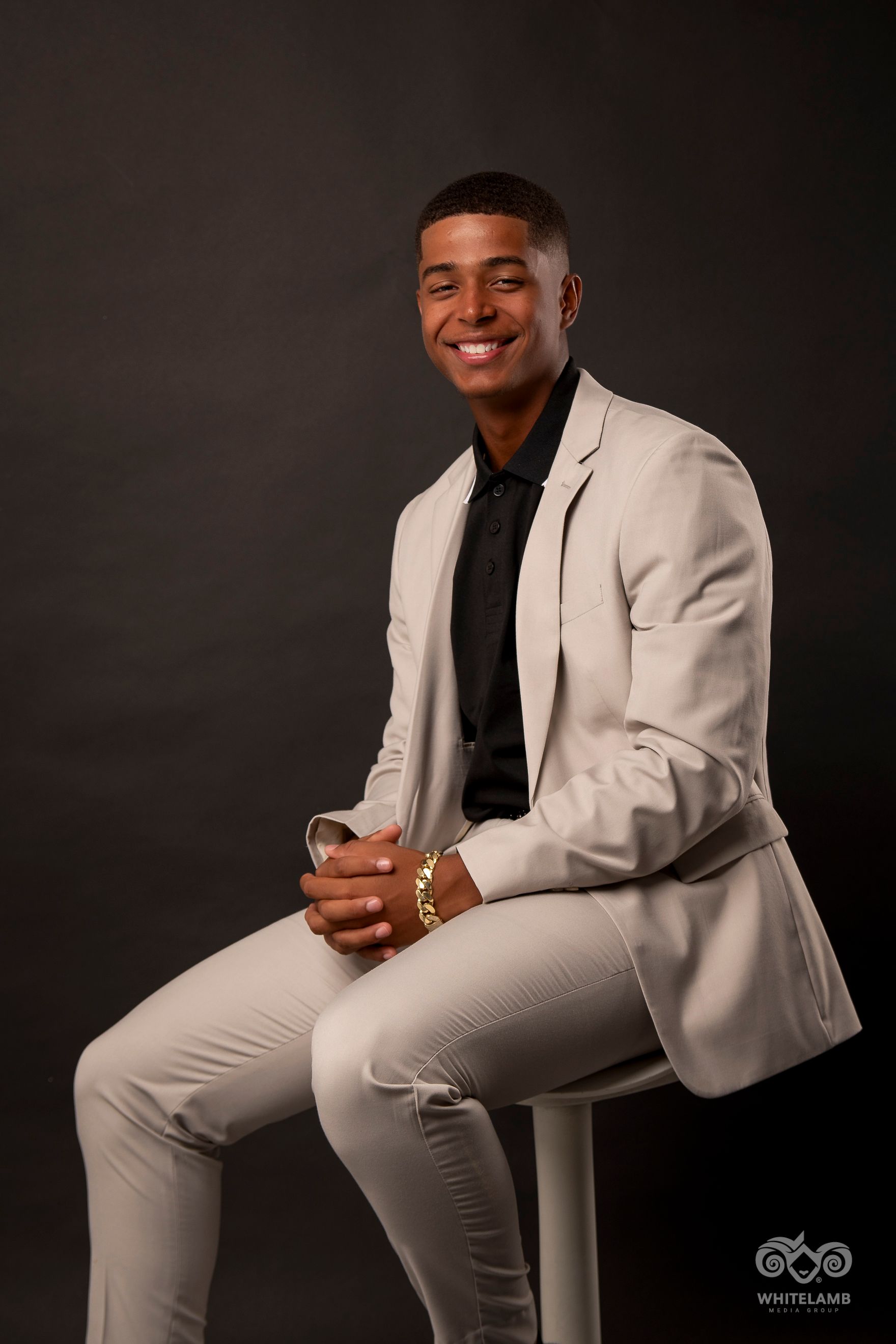 Professional studio portrait of a person wearing a light gray suit and black shirt seated on a stool against a dark background.