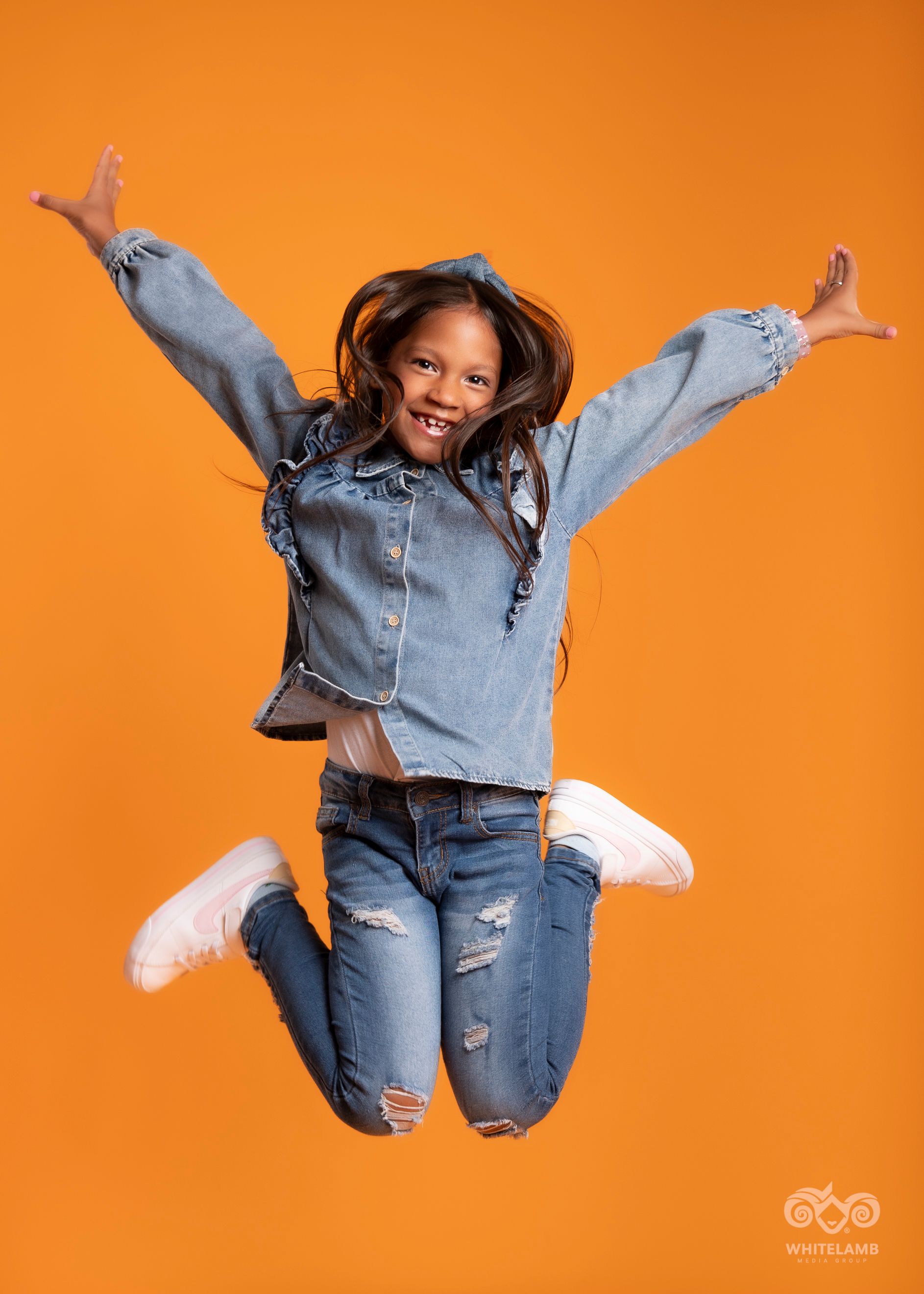 A young person wears denim coordinated outfit performs energetic jumping poses against vibrant orange background.