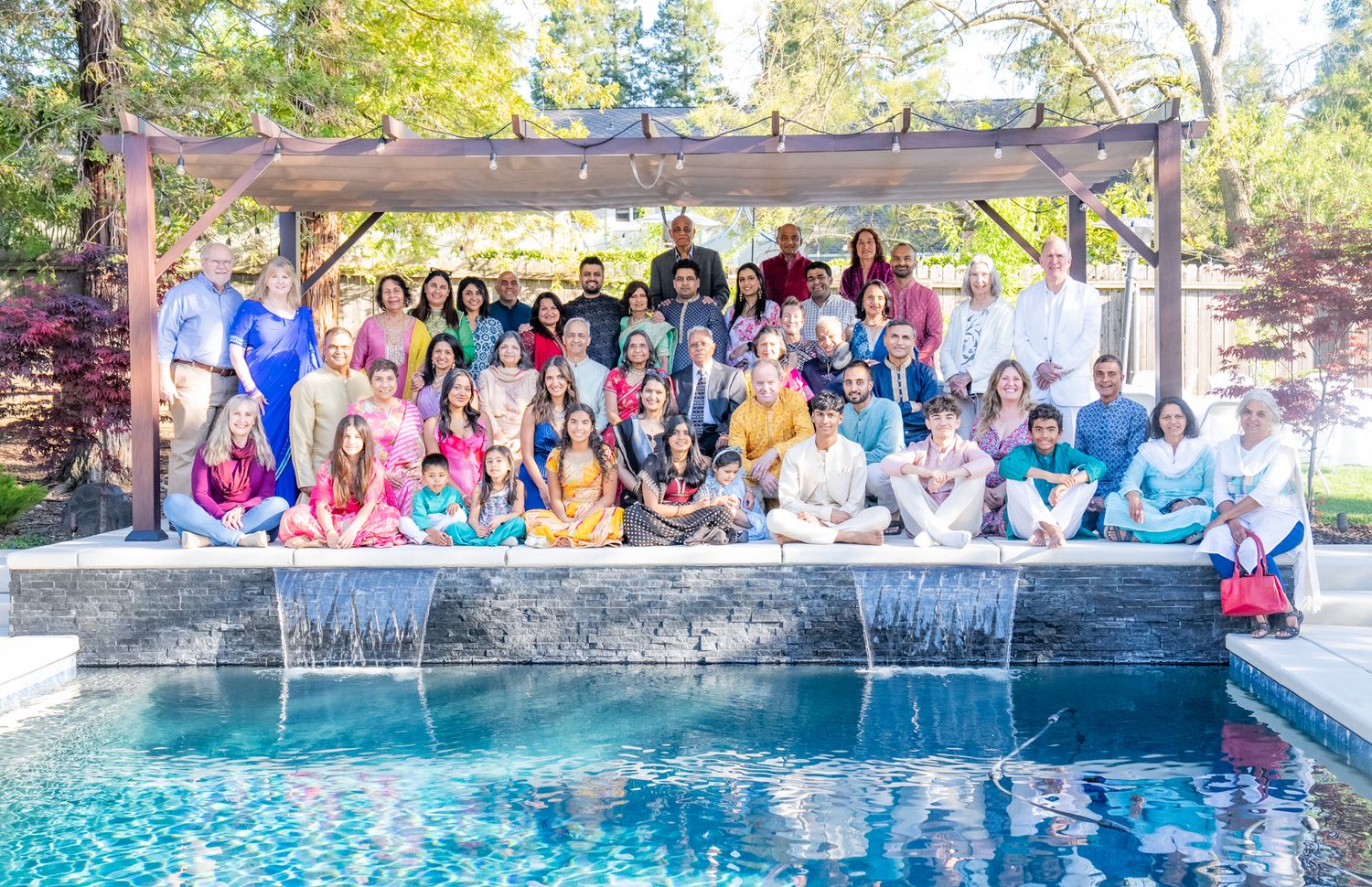 A large group gathers for a cheerful photo by a swimming pool under a wooden pergola on a sunny day.