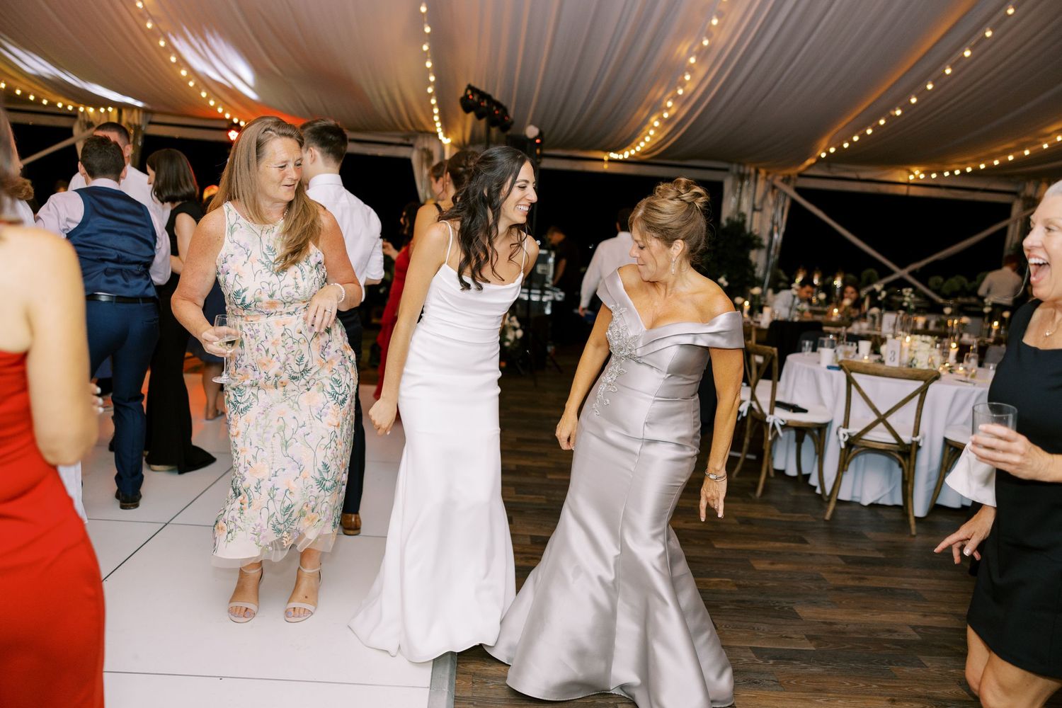 People dancing and celebrating at an elegant wedding reception under a white tent with twinkling string lights overhead.