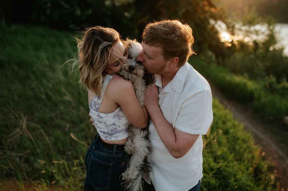 A couple shares a tender moment with their small fluffy dog at sunset on a grassy hillside path.
