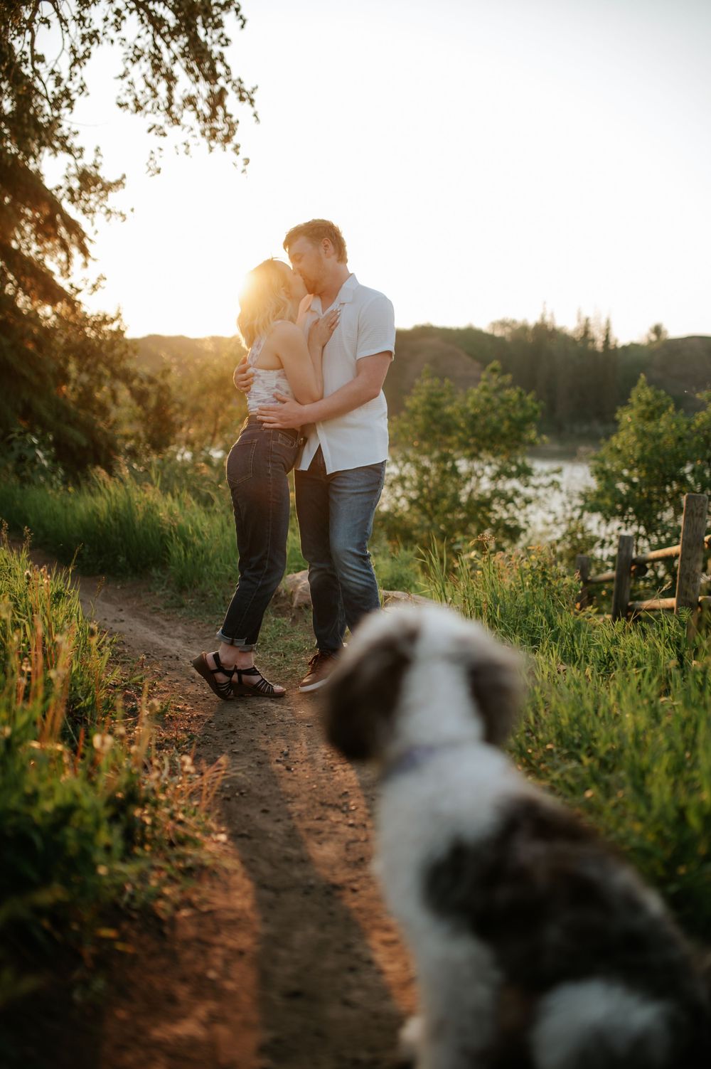 A couple shares a romantic moment at sunset on a trail path while their black and white dog watches in the foreground.