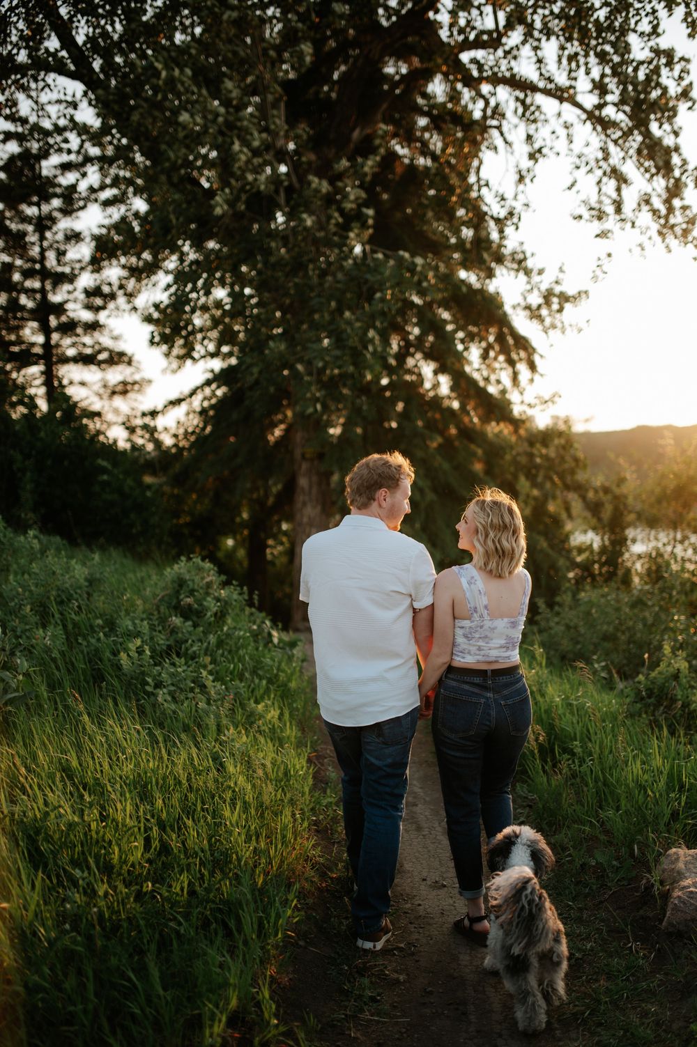 A couple and their dog walking along a scenic nature trail at sunset through tall grass and trees.