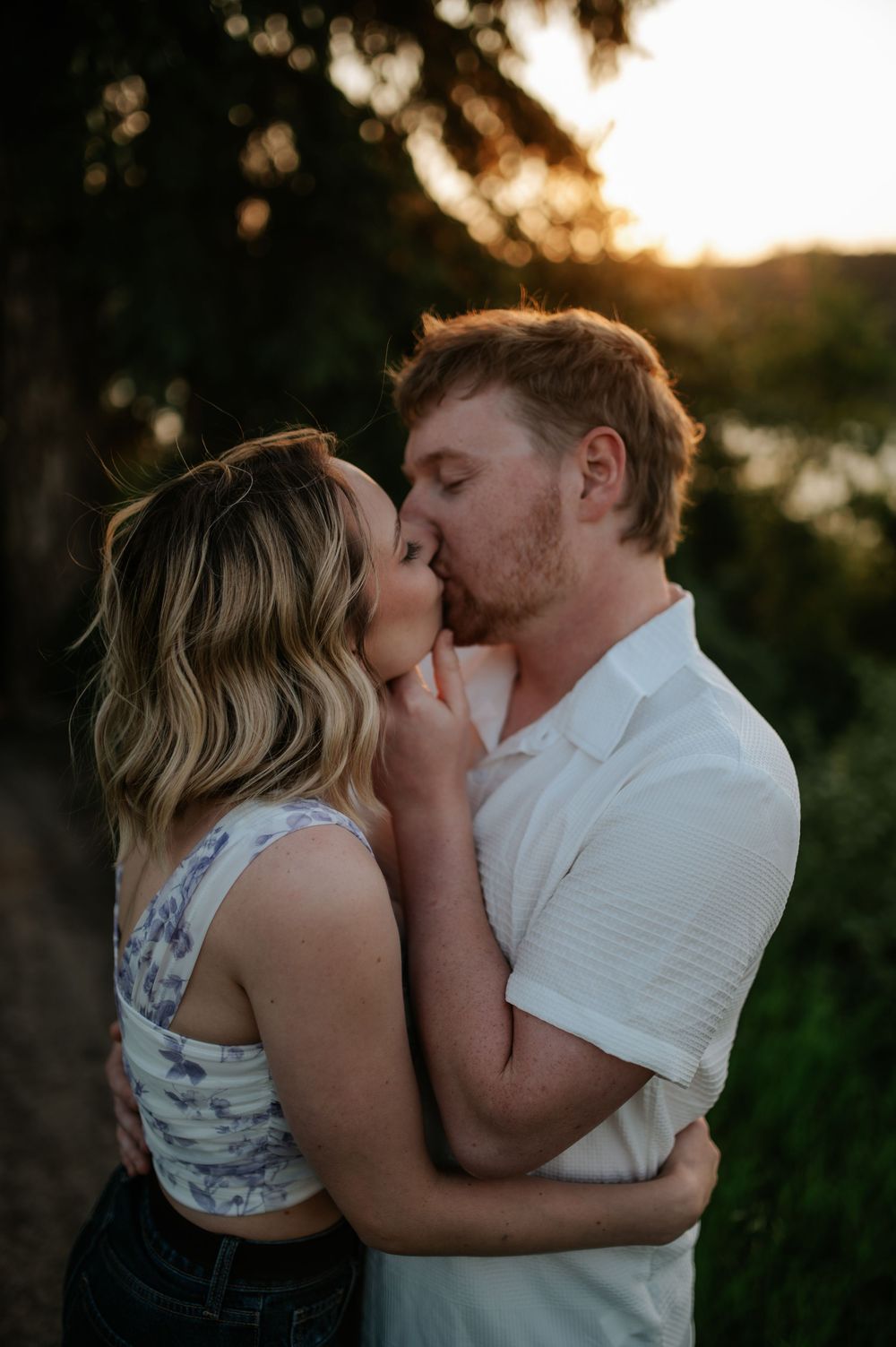 Romantic couple sharing an intimate kiss at sunset with golden bokeh light filtering through trees in the background.